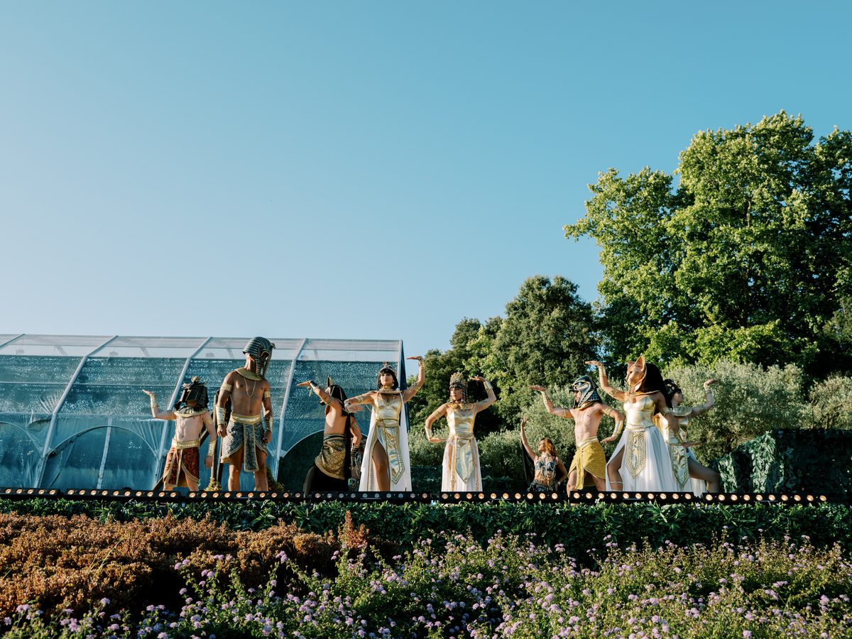 Bailarinos com trajes dourados num palco ao ar livre, num jardim ao fim da tarde